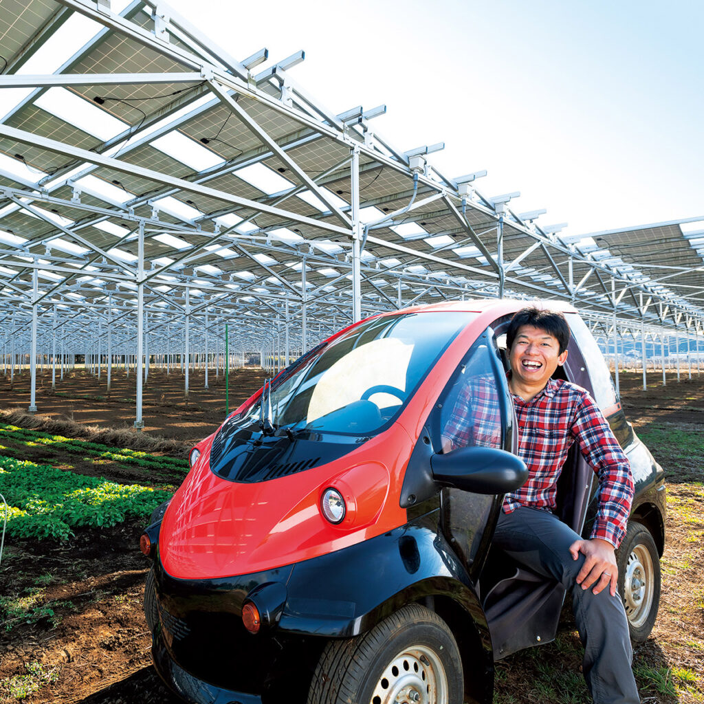 ソーラーパネルで発電されたエネルギーを充電できる社用の超小型EV(電気自動車)に乗り、笑顔を見せる馬上 丈司さん